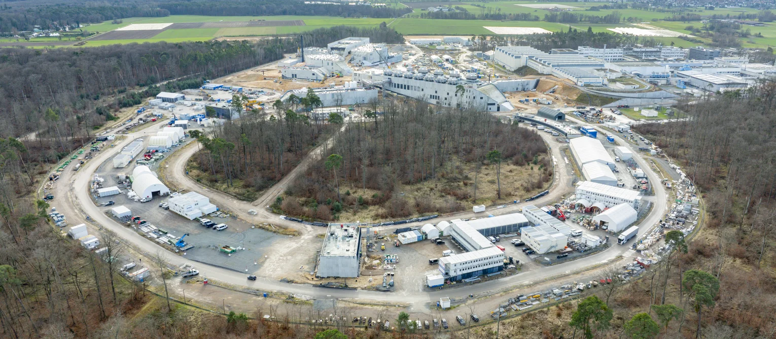 The picture shows the circular accelerator tunnel from a drone's perspective. A forest grows around the ring.