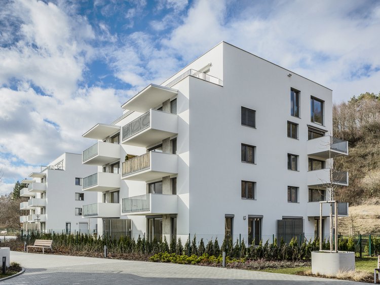 Photo: Side view Residence Pod Skalou, new building with light-coloured façade, and windows, balconies, woodland on the hill in the background, access road in the foreground