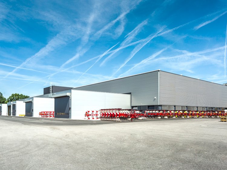 Foto: Halle mit grauer Fassade; rote landwirtschaftliche Maschinen im Vordergrund auf einer weiten Asphaltfläche