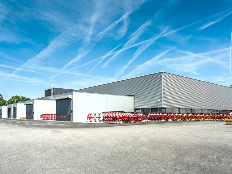 Photo: hall with grey façade; red agricultural machinery in the foreground on a broad tarmac surface