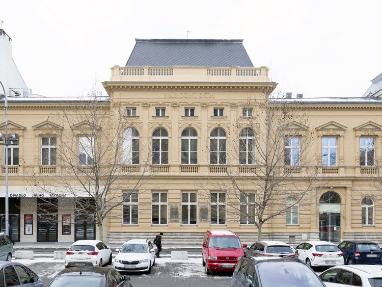 Photo: Historical building with beige facade, black roof, high windows, in the foreground parking lot with partly snowed-in cars
