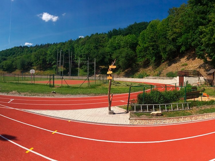 Photo: panoramic view of the stadium with sporting tracks and lawn in the middle; in the foreground, a playing field; trees in the background