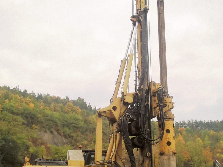 Photo: drilling apparatus on a construction site with piled-up earth and a hilly forest landscape in the background