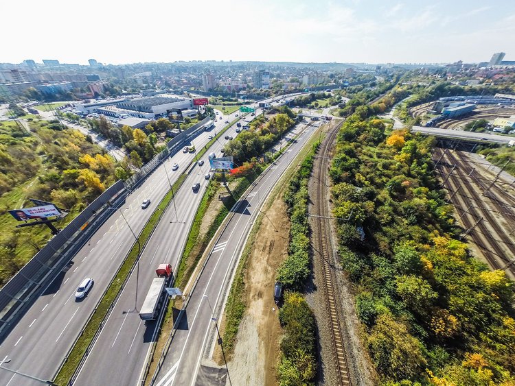 Foto: Luftaufnahme einer mehrspurigen Fahrstraβe; rechts die neue Abfahrt; weiter rechts davon die Eisenbahnstrecke umgeben von Bäumen; Großstadt im Hintergrund 