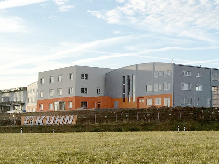 Photo: building with grey and red striped façade; in the background, a factory building with external stairs; in the foreground, a broad field 