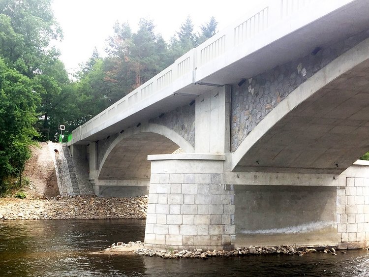Photo: view from below of a new bridge structure in historic style; underneath, a narrow river with a wooded bank on the opposite side