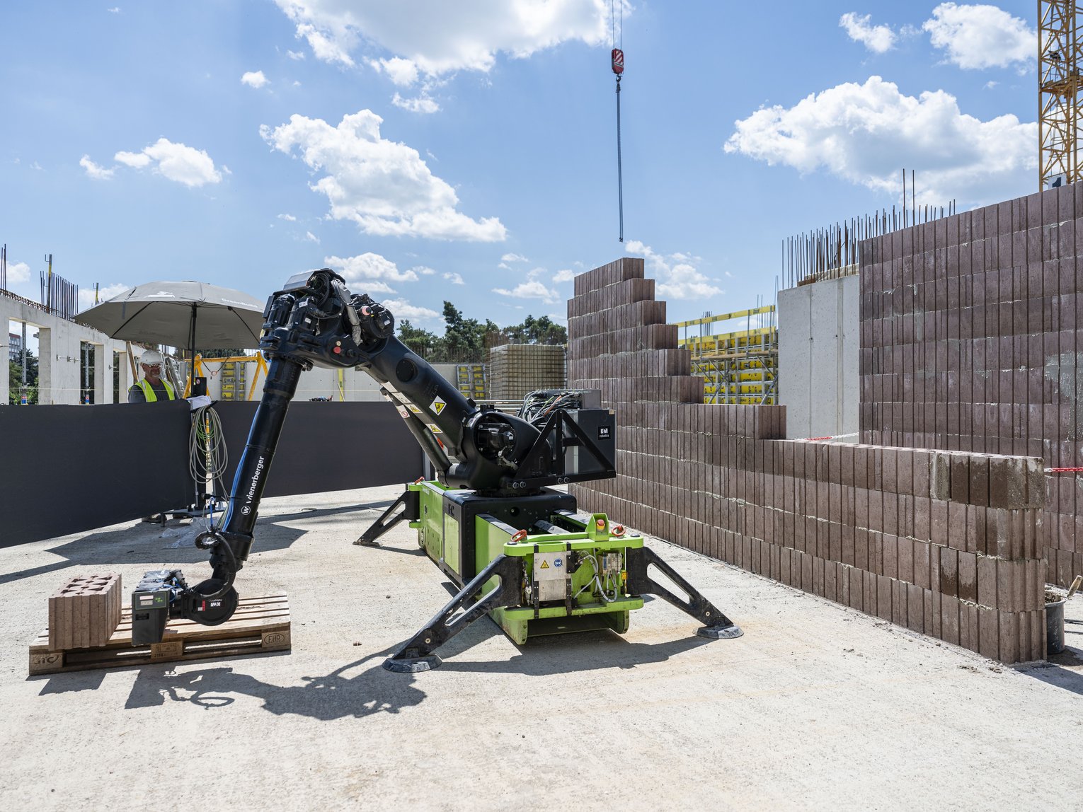 An elongated, green construction site robot with a gripper arm lifts bricks to build a wall.